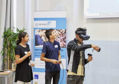 3 Kinder staunend, eines mit einer VR-Brille vor dem Werbeaufsteller von sprungbrett bayern into work